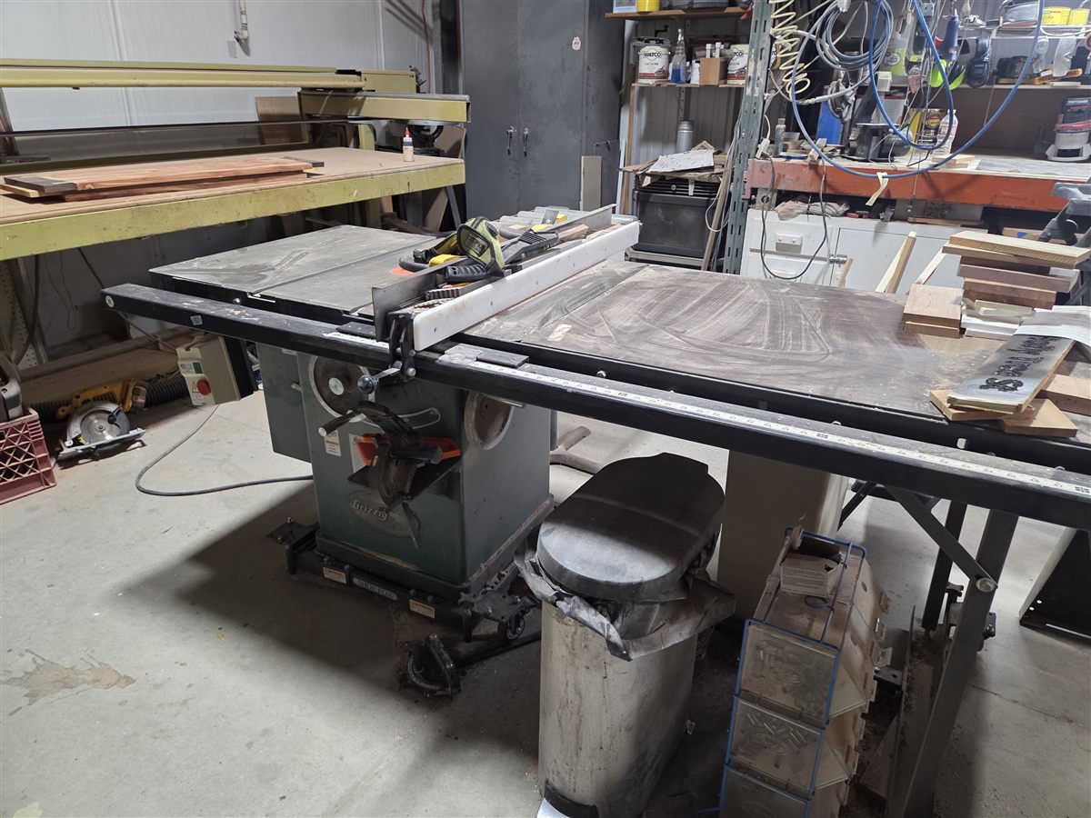 A cabinet table saw in a working woodshop, with a bench, hand tools and lumber behind it